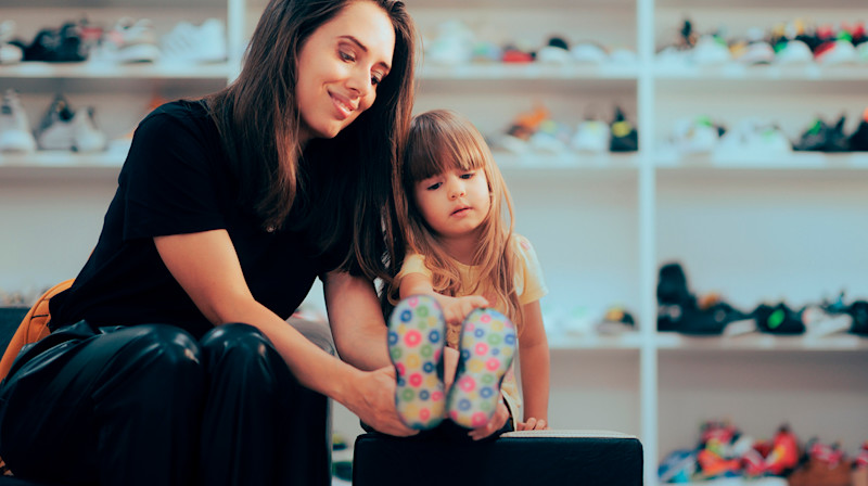 Kvinne hjelper lite barn med å prøve fargerike sko i en skobutikk med hyller fulle av sko i bakgrunnen.