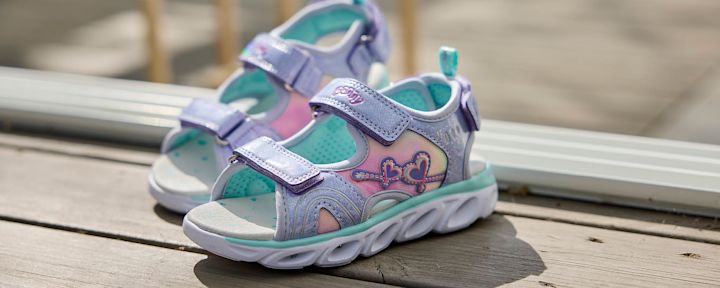 Barnesandaler i lilla og rosa med hjerte- og nøkkeldetaljer plassert på et lyst tregulv i sollys.