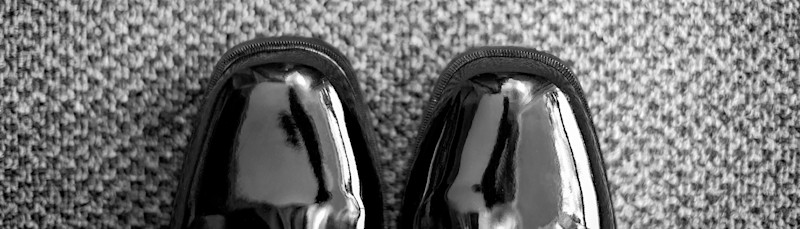 Sorte, blanke lakksko plassert på et grått, mønstret teppe sett ovenfra.